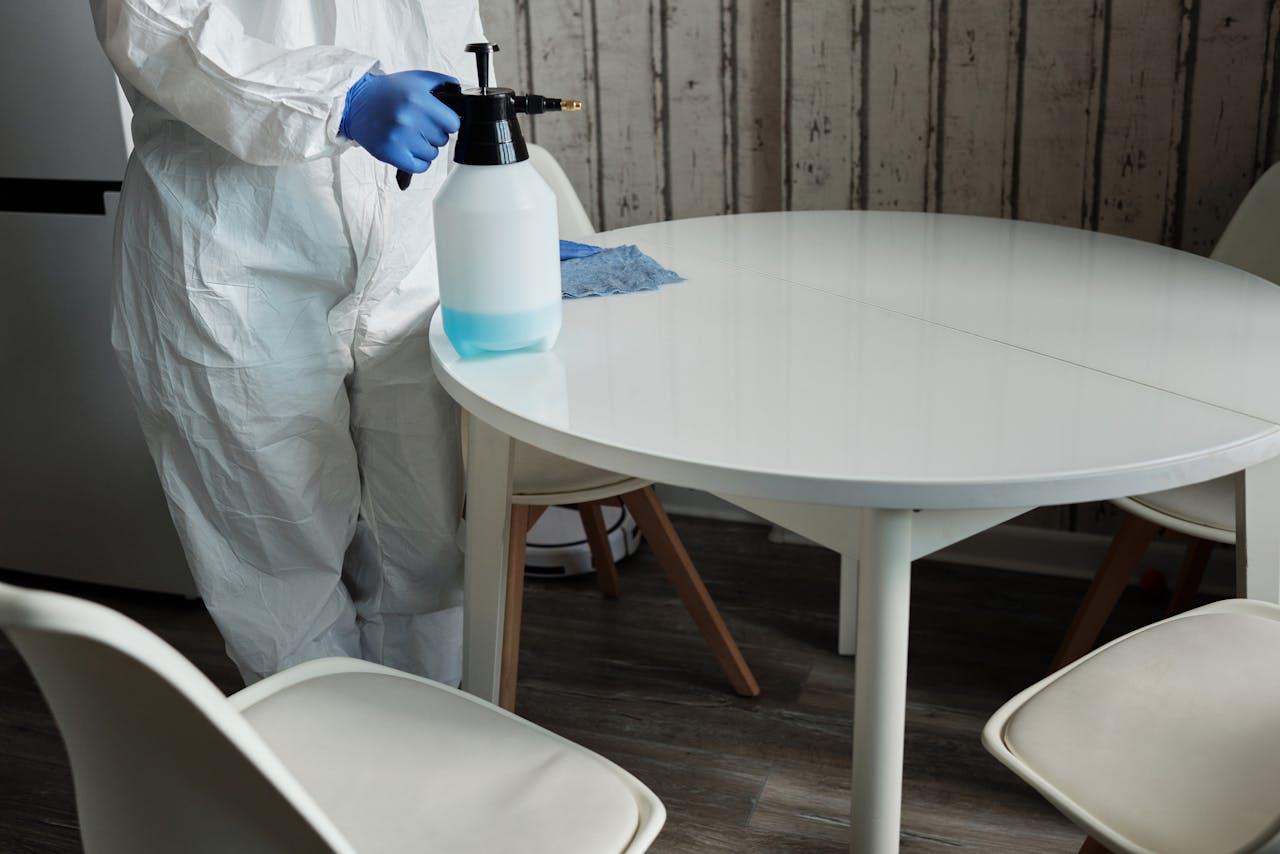 home-services-01 Person in protective suit disinfecting a white table indoors with a sprayer.